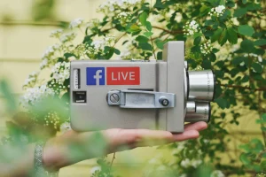 Uma mão segura uma Câmera de filmar antiga com autocolante de facebook live, num fundo de verdes plantas, tal como na gestão de redes sociais.